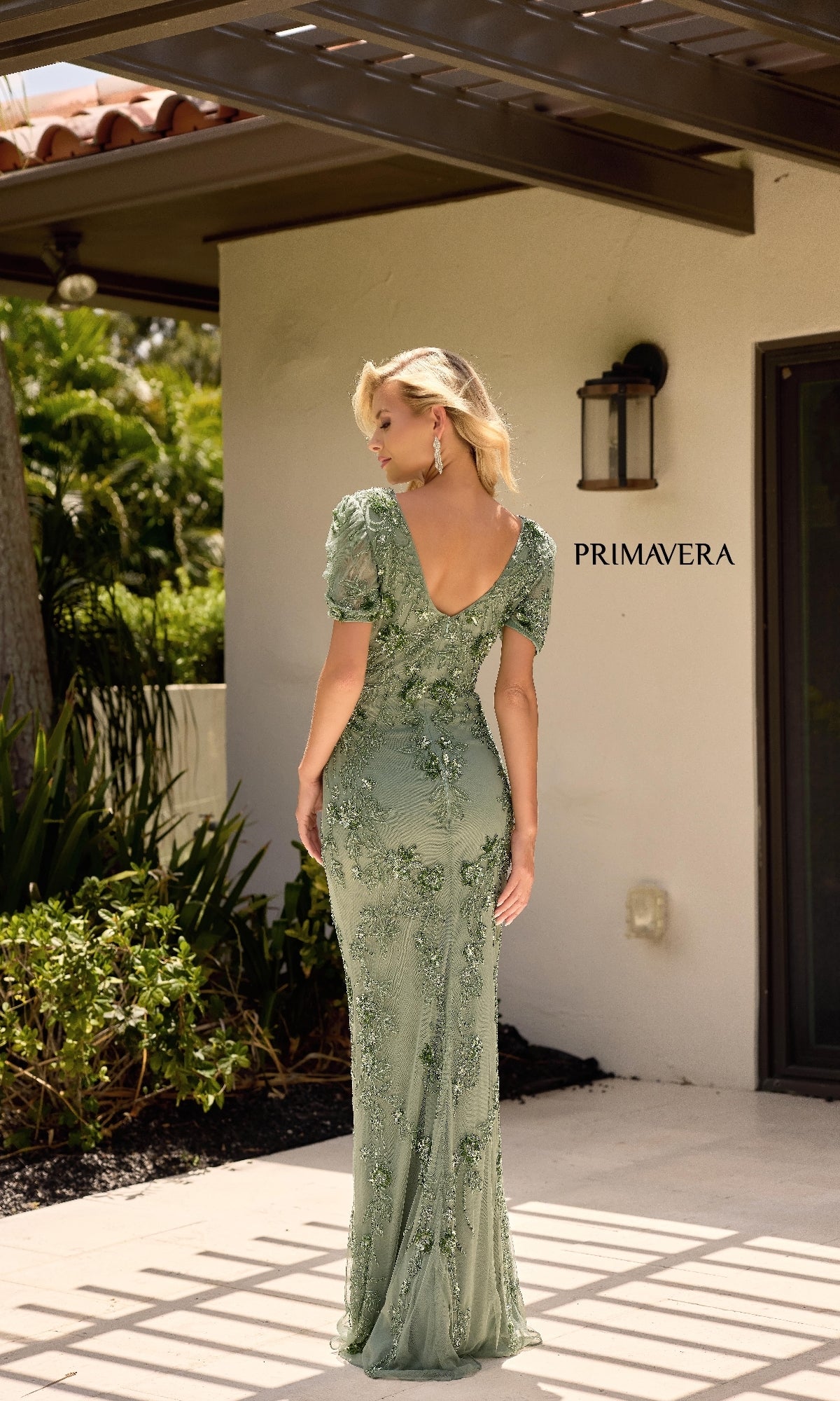 A woman stands outdoors facing away, wearing the Primavera 13147 long formal dress in light green with short sleeves, floor-length style, and a beaded floral design.